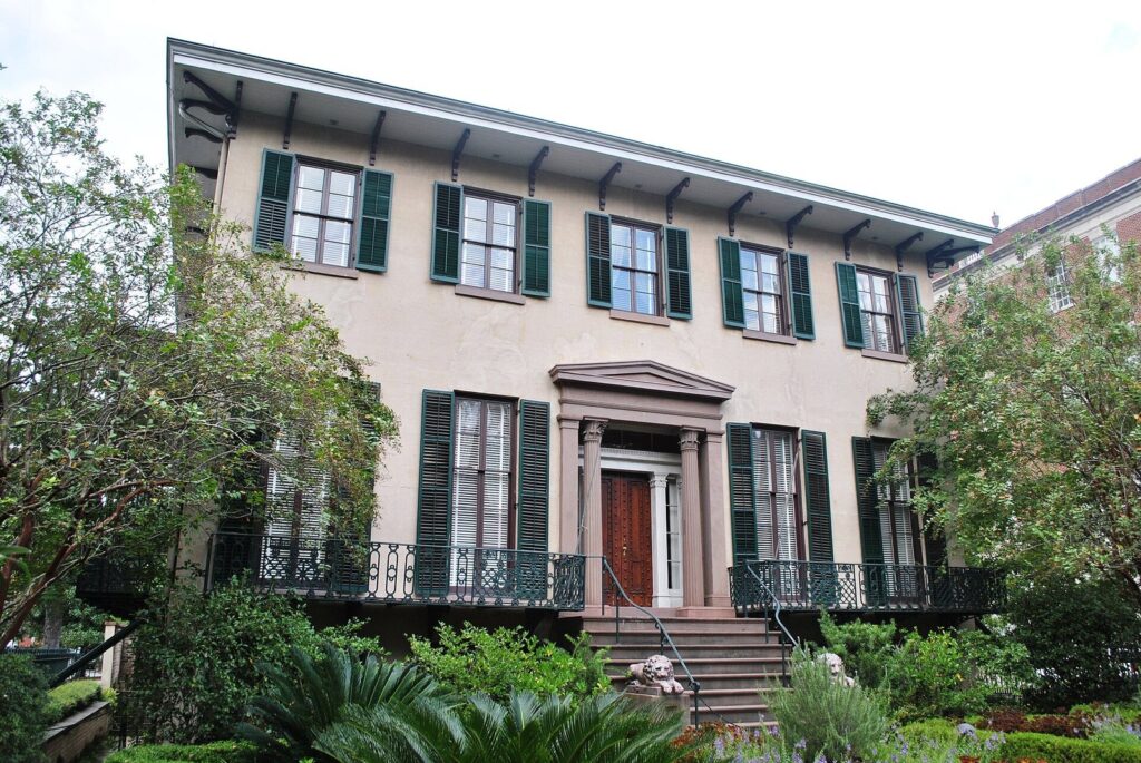 The stucco front of the Andrew Low House in Savannah, with green shutters, iron balconies and a garden in front