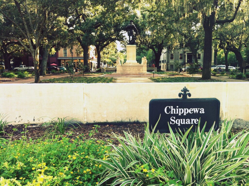 The sign for Chippewa Square in a planted bed, with the square's shaded paths and the Oglethorpe statue behind