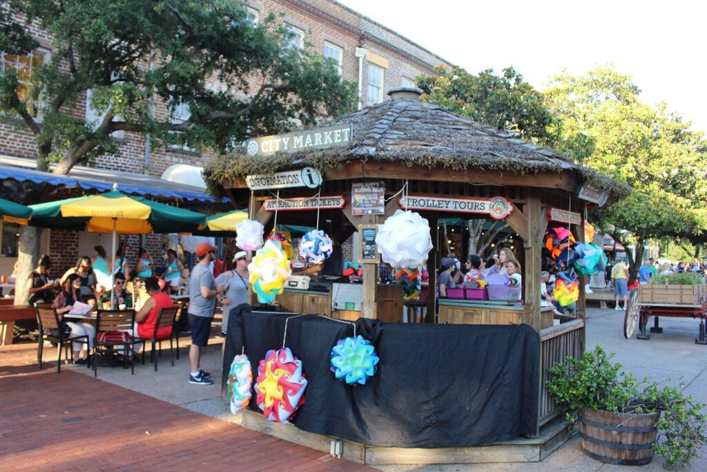A thatched information booth selling trolley tours in City Market, Savannah, with diners at umbrella tables around it