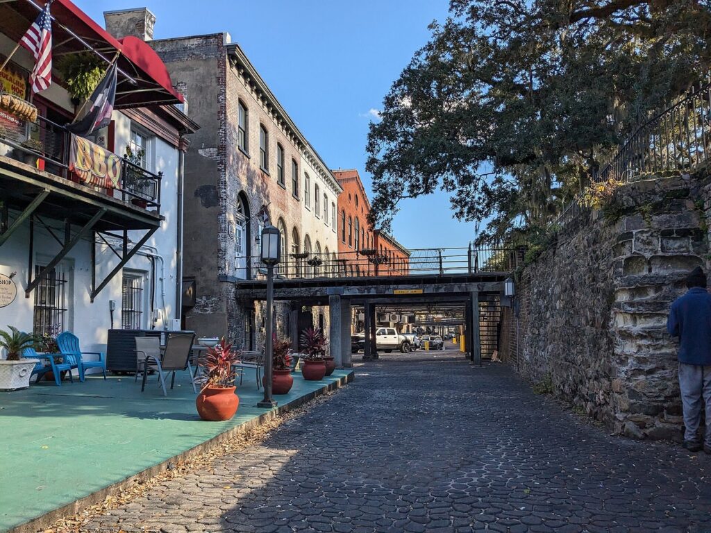 The brick buildings and stone retaining wall of Factors Walk in Savannah, with an iron bridge over the lane