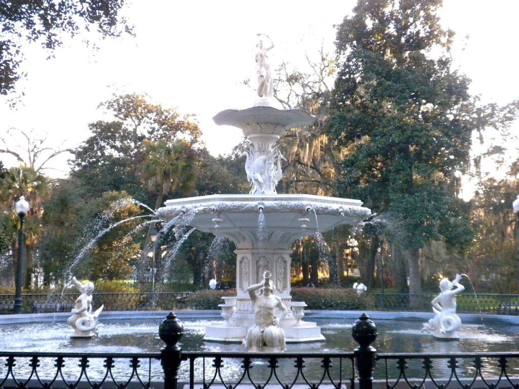 The white Forsyth Park fountain sending arcs of water into its basin, framed by live oaks and palms