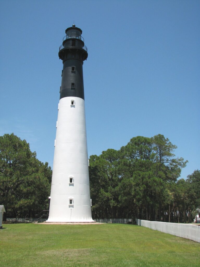 The tall white and black Hunting Island Lighthouse rising above pine trees under a blue sky