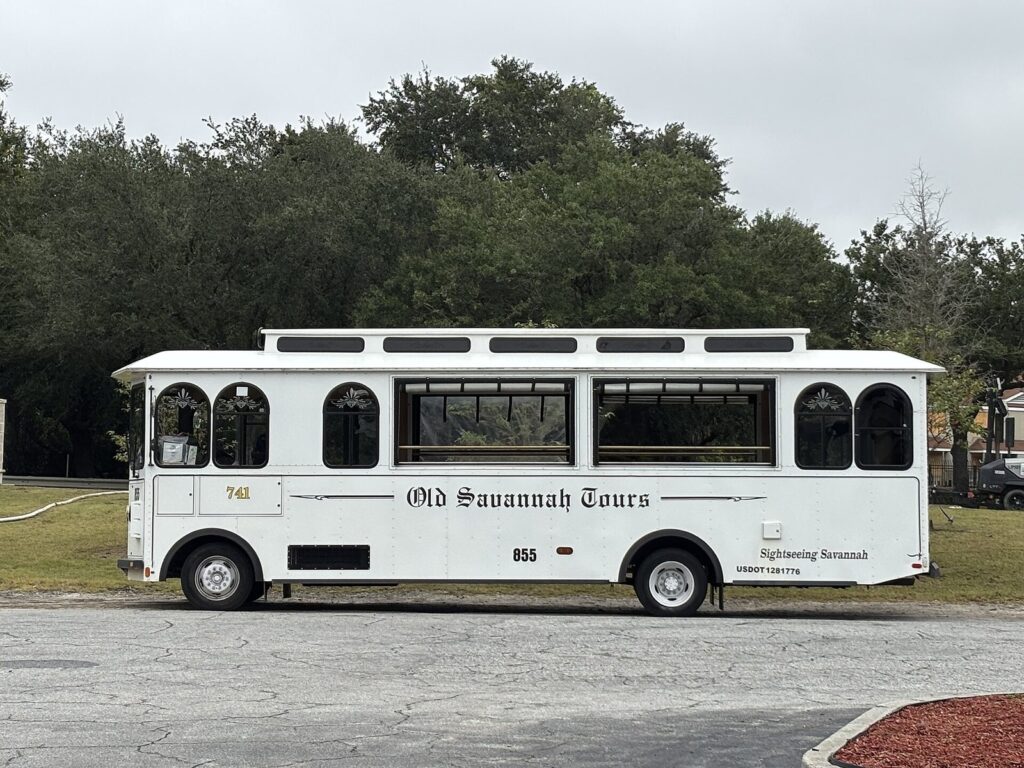 A white Old Savannah Tours trolley bus parked beside trees in Savannah