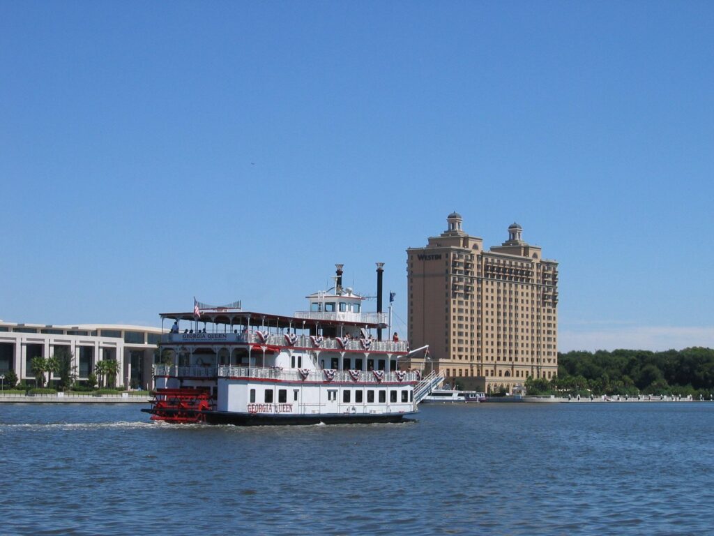 A white paddlewheel riverboat cruising the Savannah River with a tall hotel on Hutchinson Island behind it