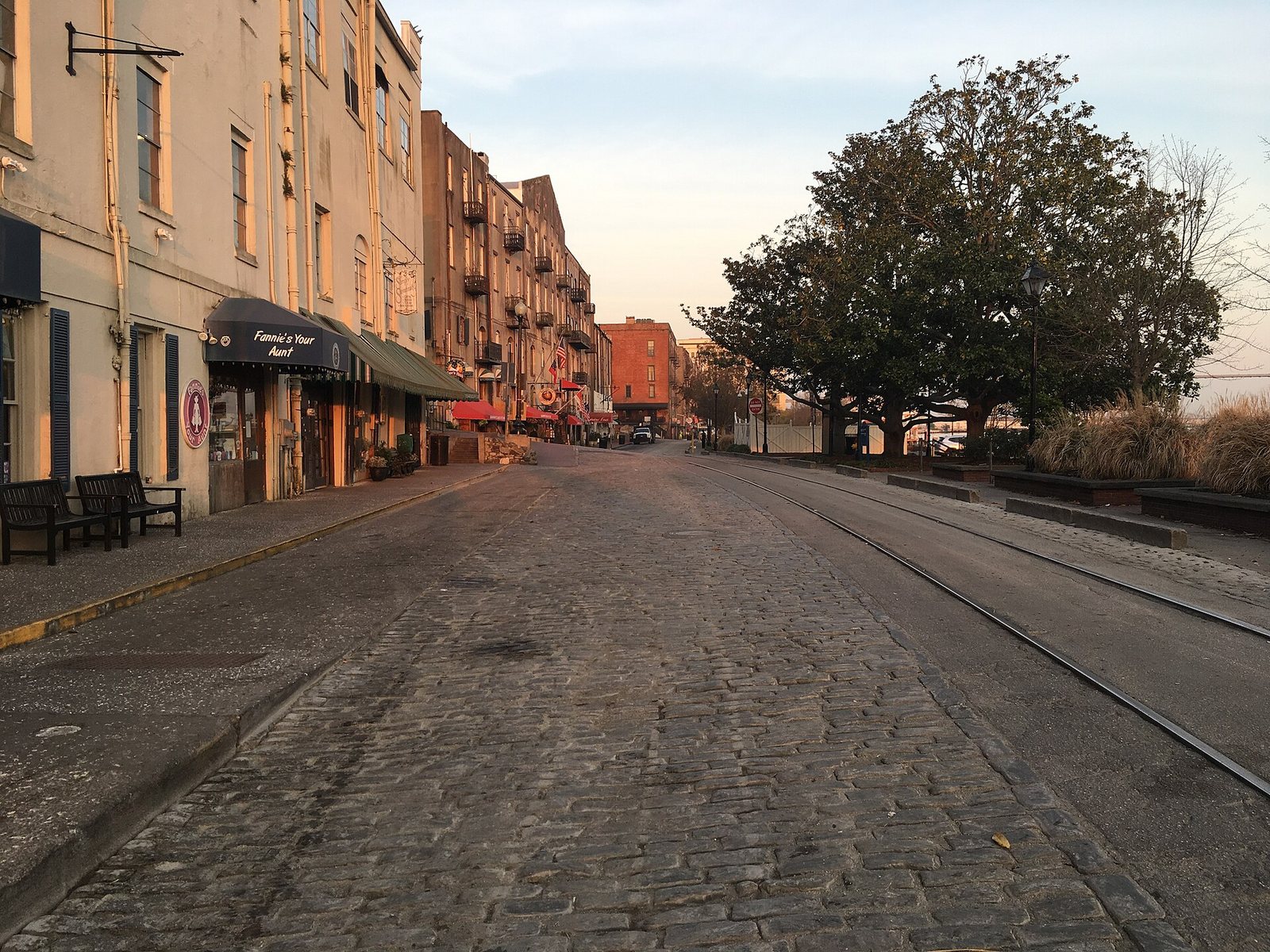 Empty cobblestone River Street at dawn with old brick cotton warehouses converted to shops on the left and trees along the river on the right