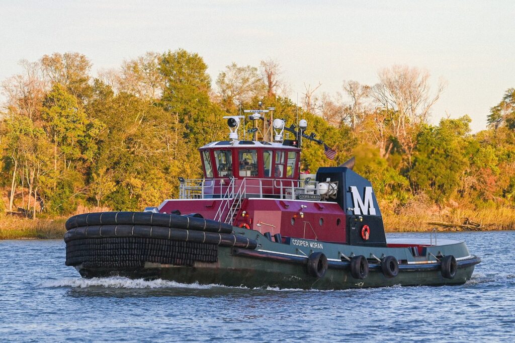 A red and black Moran tugboat pushing through the Savannah River past marsh and trees in autumn light
