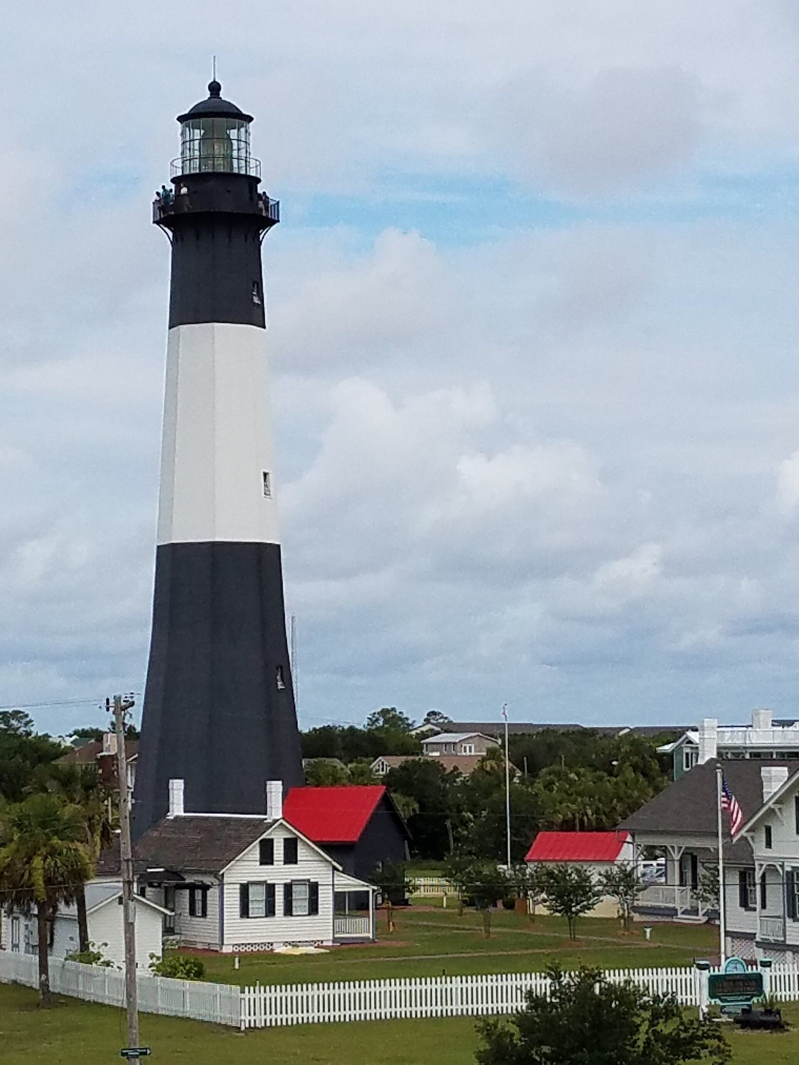 The black-and-white Tybee Island lighthouse tower above white keeper cottages with red roofs behind a picket fence