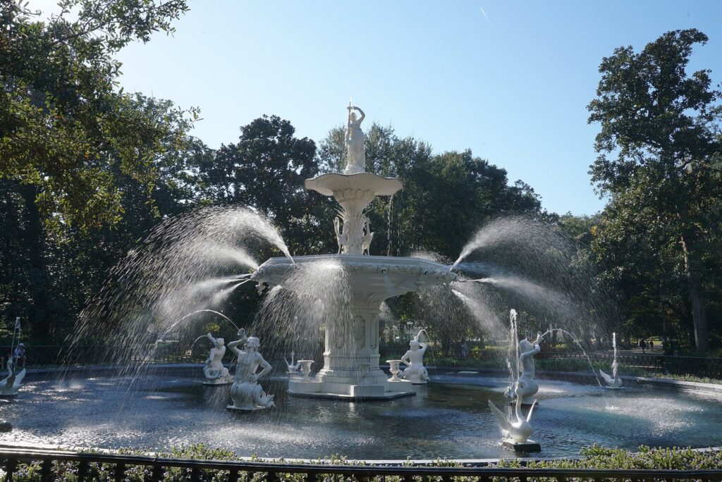 The white cast-iron Forsyth Park fountain spraying water in tiers beneath a blue sky, surrounded by an iron fence and live oaks