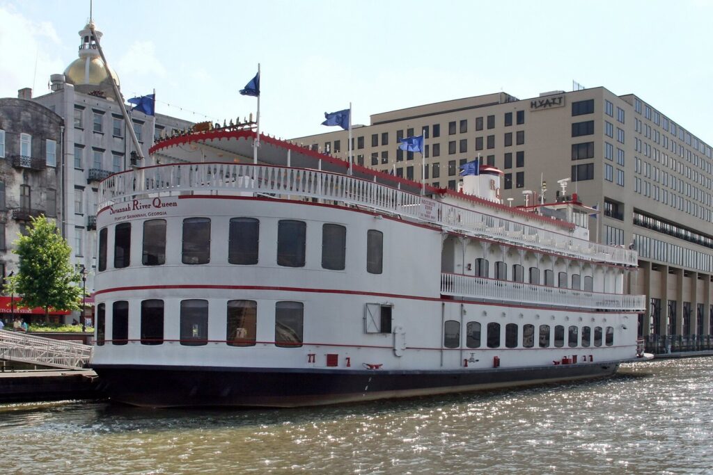A white riverboat with red trim docked on the Savannah River below City Hall's gold dome and the Hyatt Regency